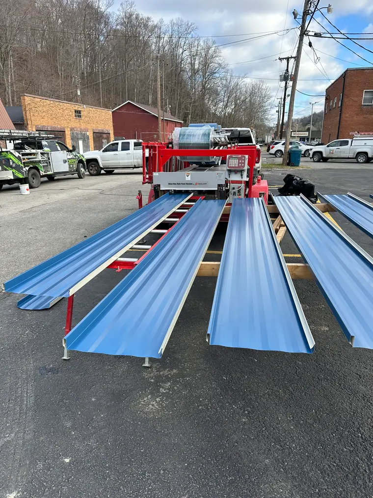 Portable roll-forming machine on a trailer producing long blue metal roofing panels in a parking lot, work trucks parked nearby