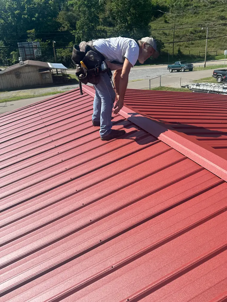 Roofer wearing a tool belt fastening a red metal roof, with the town and parked trucks visible below