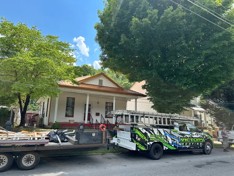 Victory Roofing branded truck and flatbed trailer parked in front of a white home with a new clay metal roof, crew member at the curb