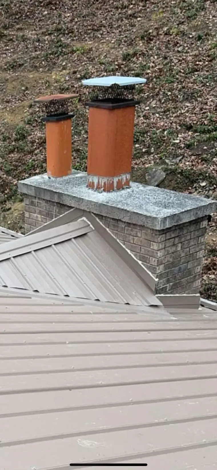 Two capped clay-colored chimney flues rising from a clay standing-seam metal roof with a wooded hillside behind