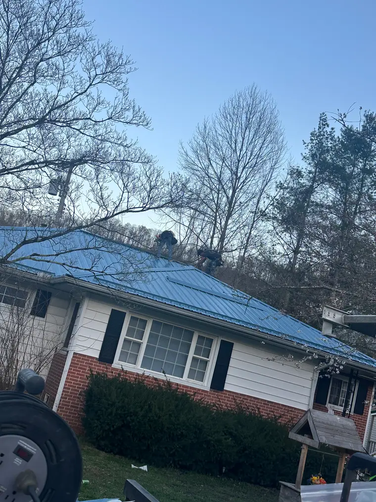 Crew installing a blue metal roof on a white-sided cottage with a brick base at dusk, bare trees around the property
