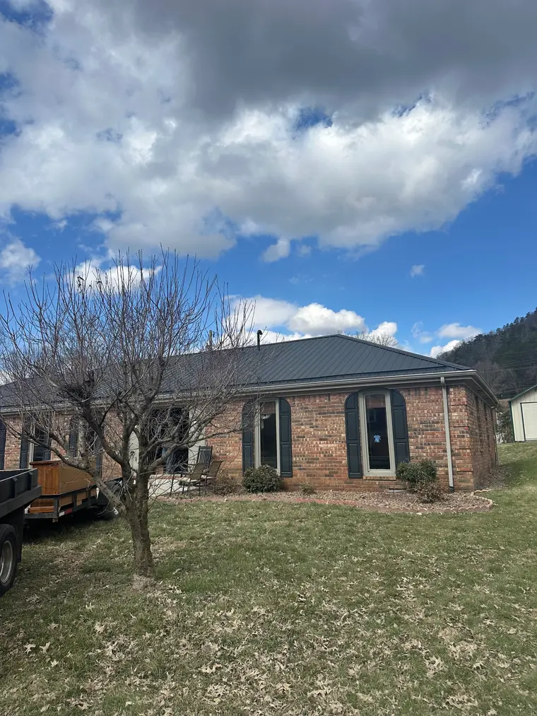 Brick ranch home with a dark bronze metal hip roof, a bare tree in the front yard and a wooded hillside behind