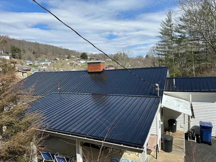 Overhead view of a black standing-seam metal roof with a brick chimney on a hillside home, neighborhood and bare trees beyond