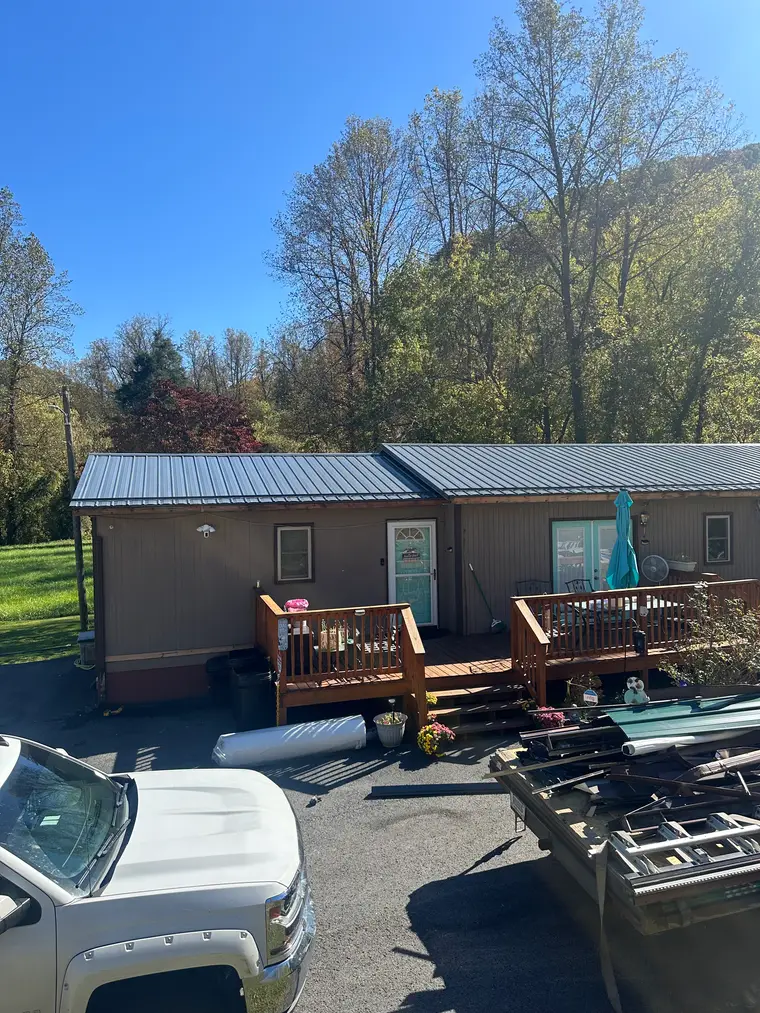 Brown modular home with a black metal roof and a wooden covered deck on an autumn hillside, with a work truck and materials in the foreground
