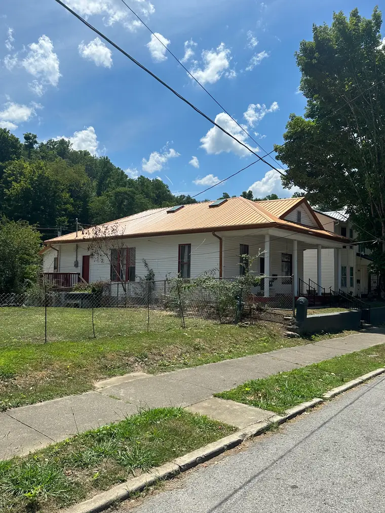 White historic farmhouse with a wrap-around porch and a clay-colored standing-seam metal roof, viewed from the street behind a chain-link fence
