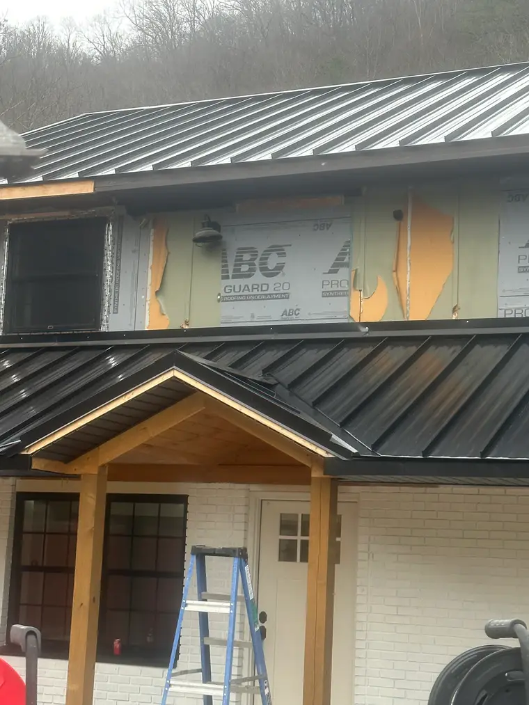Two-story mountainside home being re-roofed with black standing-seam metal panels over a cedar-post porch, with synthetic underlayment exposed on the upper wall