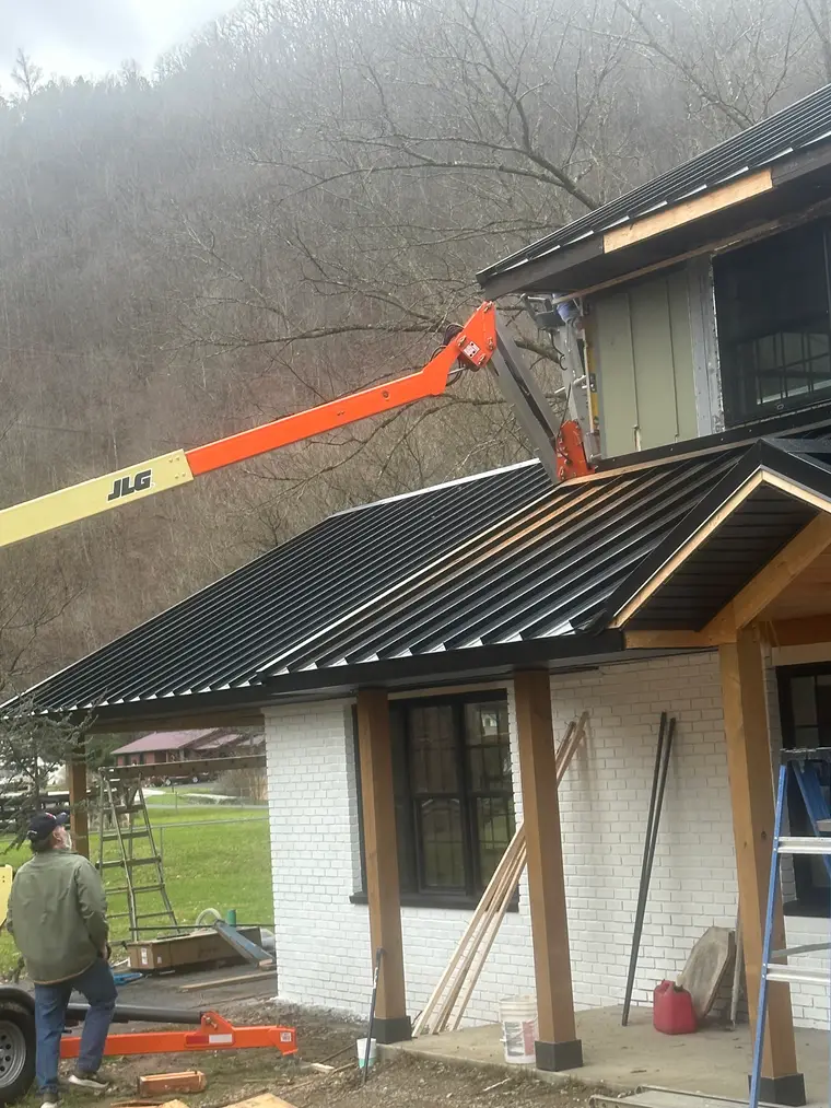 Roofer on an orange lift installing a black standing-seam metal roof over the white-brick porch of a mountainside home
