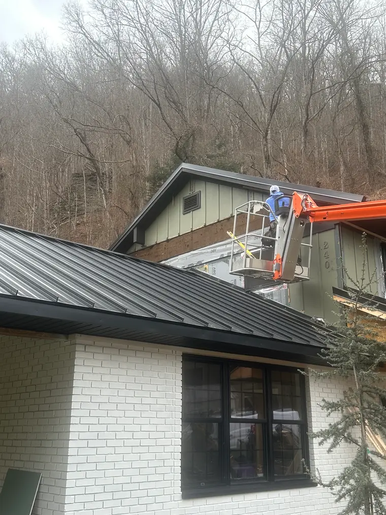 Wide view of a white-brick home on a mountainside with a black standing-seam metal roof and a worker on a lift, bare winter trees behind