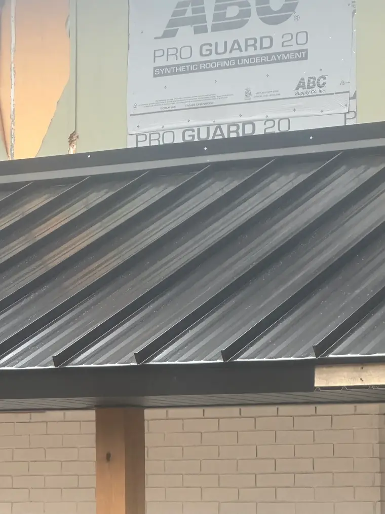 Detail of black standing-seam metal roof panels and trim with roofing underlayment visible on the wall above a brick column