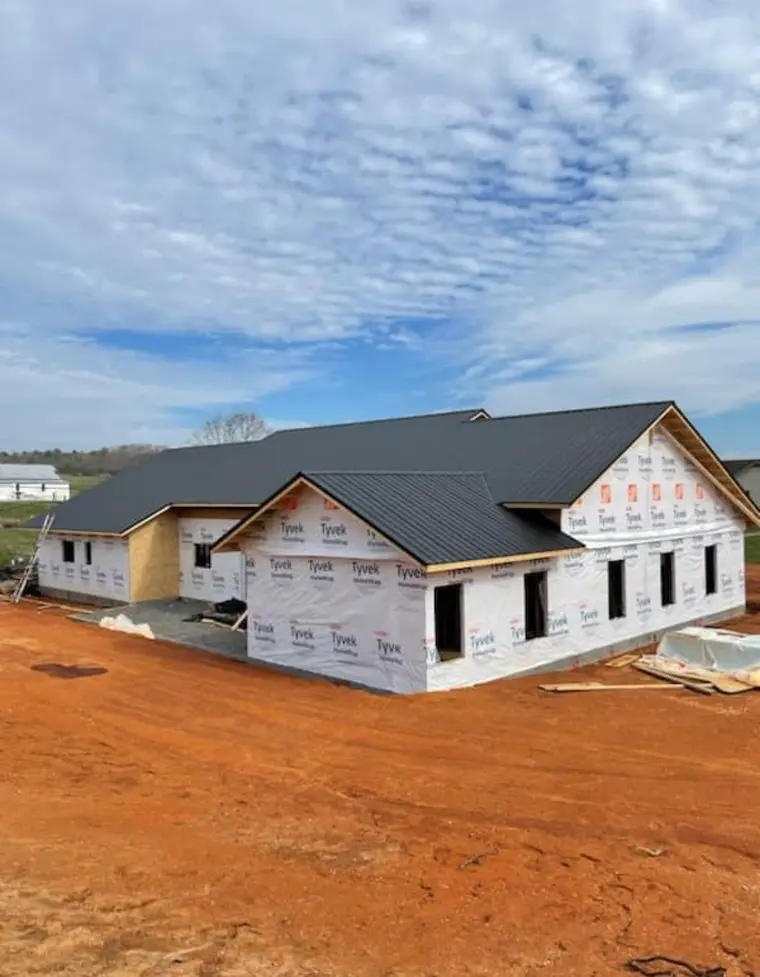 Newly framed ranch home wrapped in house wrap with a freshly installed black metal roof, surrounded by red-dirt construction yard