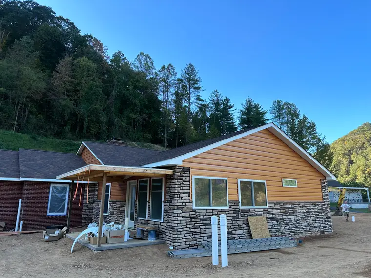 Single-story home with cedar-tone lap siding, stacked-stone veneer and a brick wing, covered porch and mountainside behind