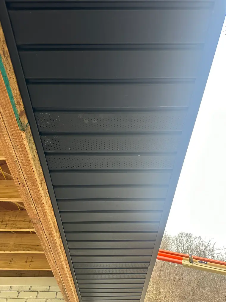 Black metal soffit with integrated venting installed beneath a roof eave, wood framing visible at the edge